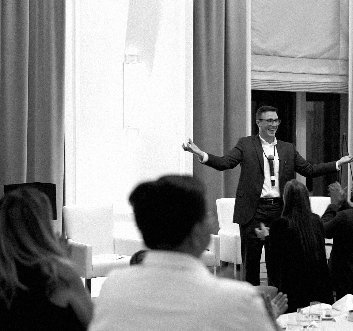 From the back of a banquet room, guests at dinner tables applaud a performer at the front.