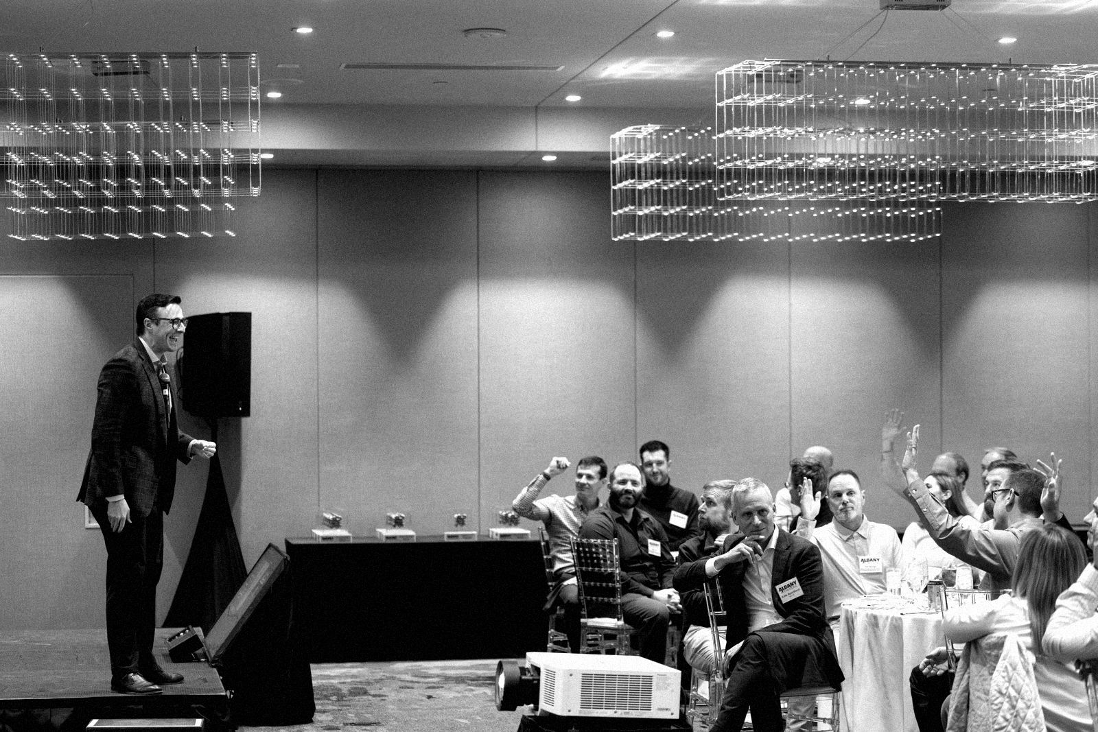 A corporate audience at round tables raising their hands as a performer laughs from a low stage.